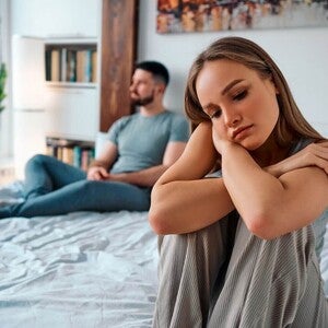 Fotografija tužne žene na rubu kreveta, partner udaljen u pozadini
