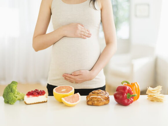 Trudnica stoji ispred stola sa voćem, povrćem i kolačima i drži se za trbuh;