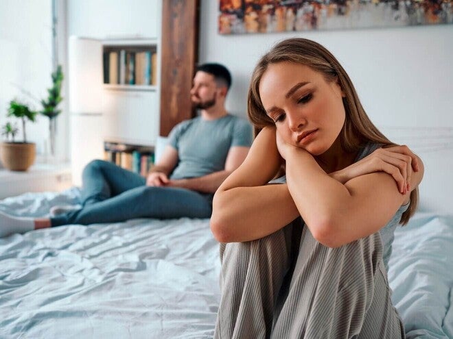 Fotografija tužne žene na rubu kreveta, partner udaljen u pozadini