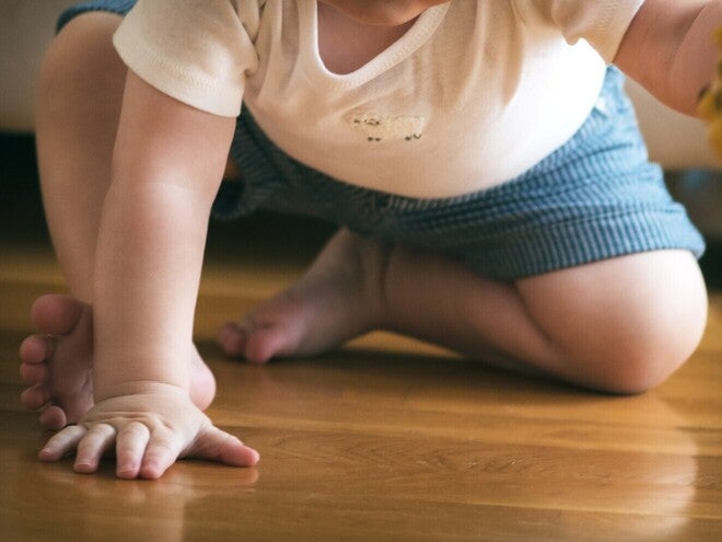 Fotografija bebe koja puzi po drvenom podu, u beloj majici i plavim šorcima, drži žutu igračku.