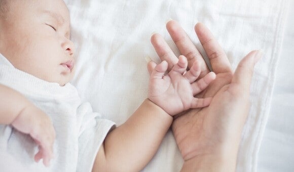 Fotografija spavajuće bebe koja drži odraslu ruku na belom plahtu