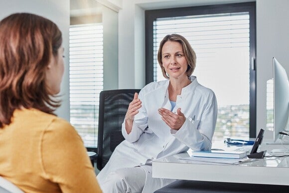 Photograph: female doctor in white coat smiling and gesturing to seated patient in bright office.