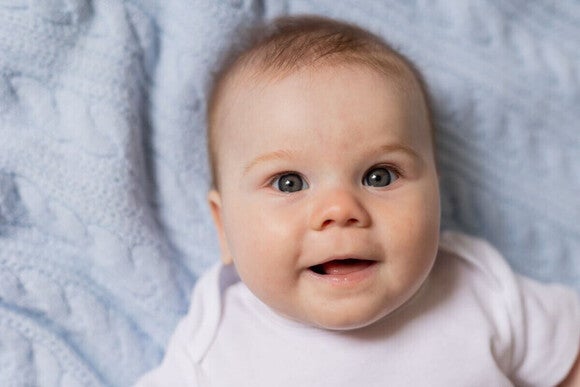 Smiling baby on light blue knitted blanket, wearing a white onesie.